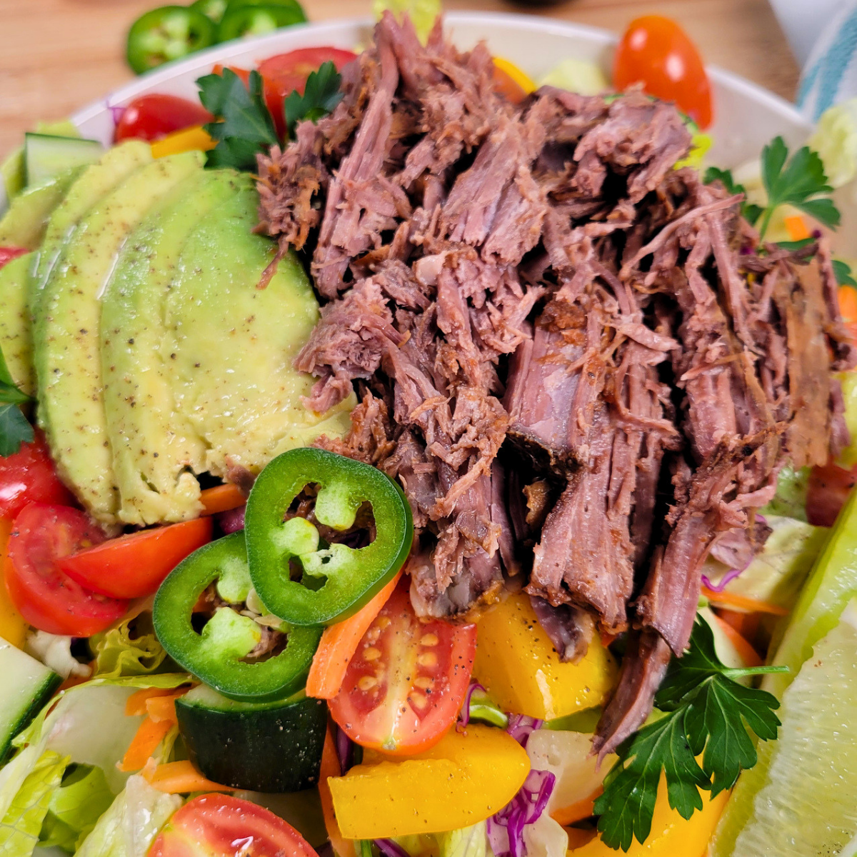 A colorful salad topped with shredded beef, sliced avocado, jalapeño, cherry tomatoes, cucumber, yellow bell peppers, shredded carrots, and parsley on a bed of leafy greens.