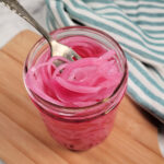 A fork lifts bright pink pickled onions from a glass jar on a wooden board, with a blue and white striped cloth in the background.