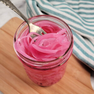 A fork lifts bright pink pickled onions from a glass jar on a wooden board, with a blue and white striped cloth in the background.