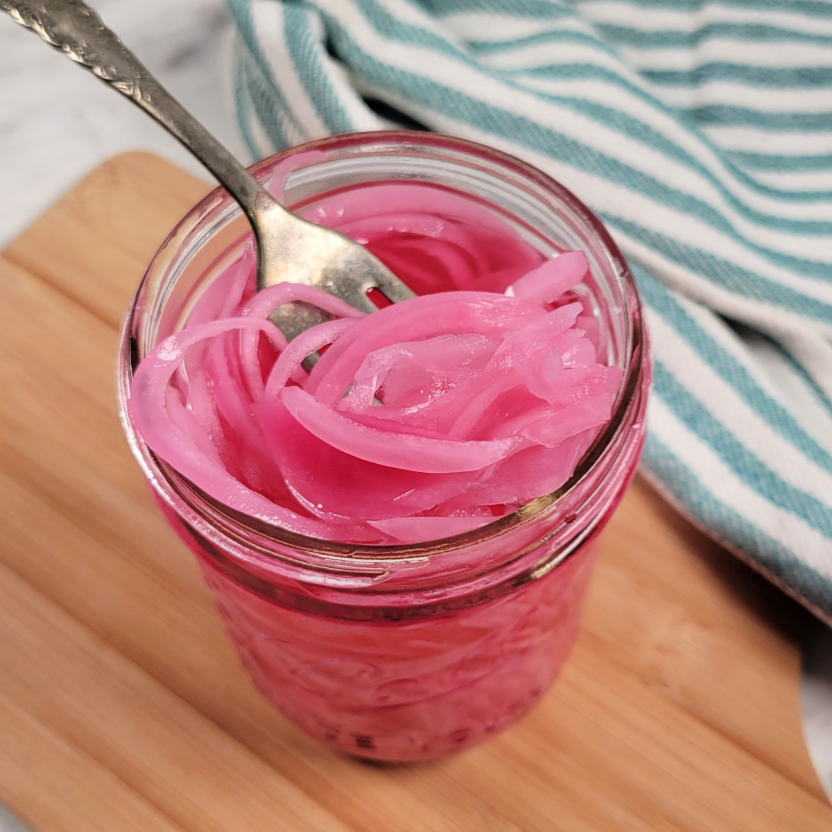 A fork lifts bright pink pickled onions from a glass jar on a wooden board, with a blue and white striped cloth in the background.