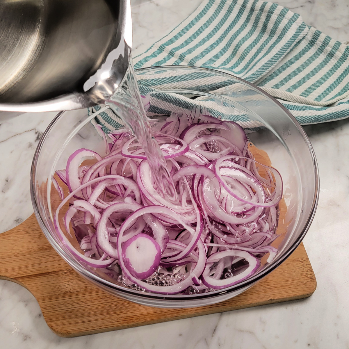 A hand pours water from a metal pot over sliced red onions in a glass bowl on a wooden board, with a striped kitchen towel in the background on a marble countertop.