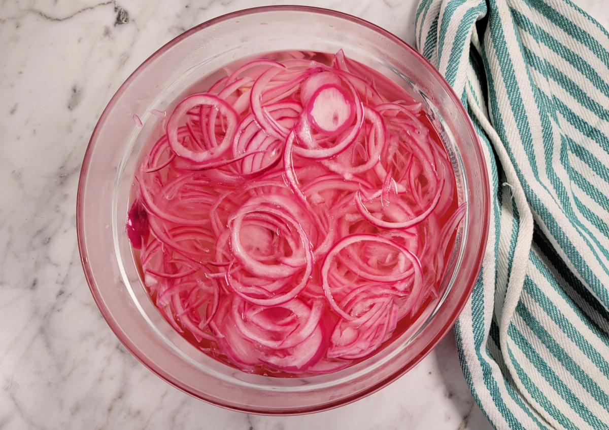 A glass bowl filled with thinly sliced red onions soaking in water sits on a marble countertop next to a green and white striped towel.