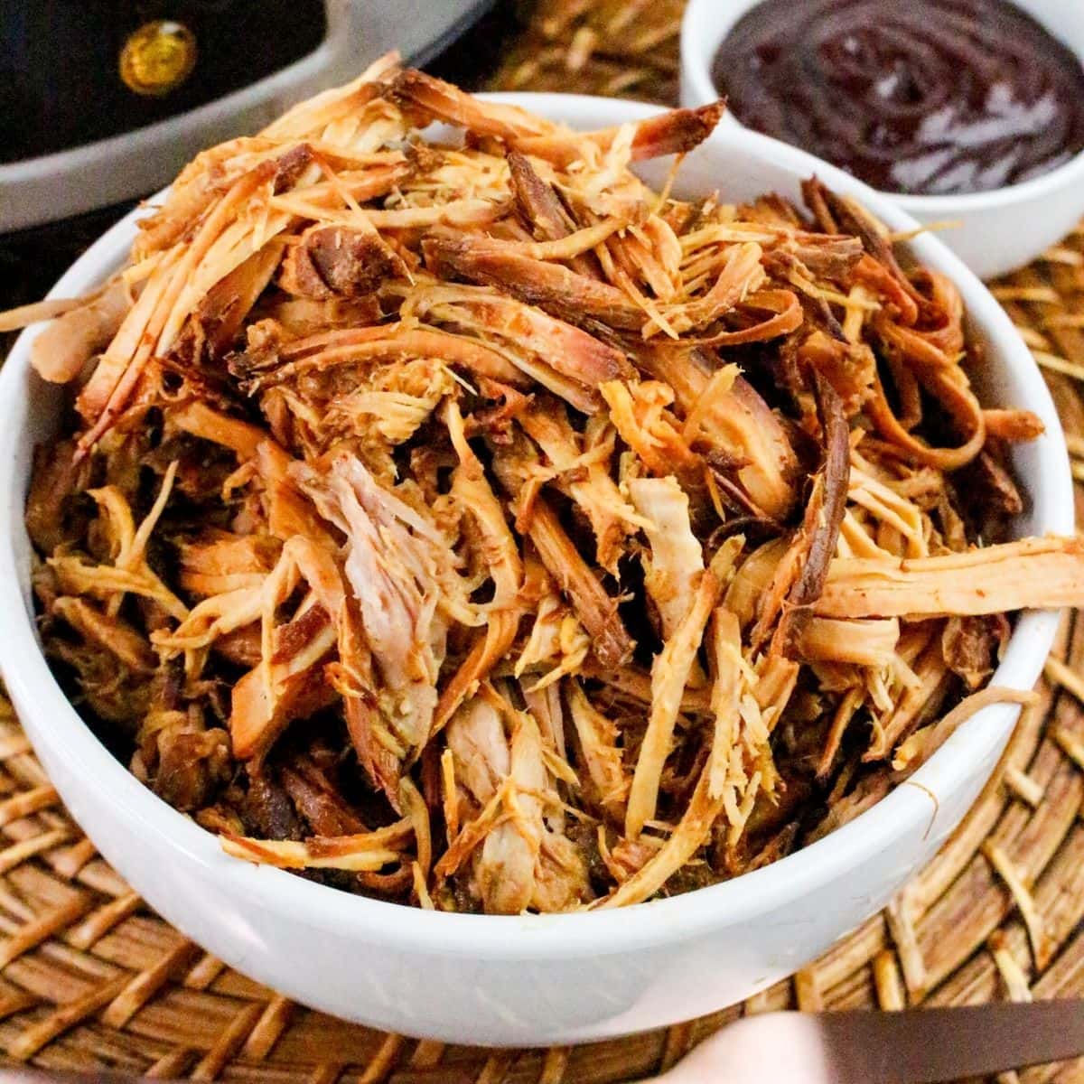 Crockpot BBQ Pulled Pork: a white bowl filled with shredded pulled pork sits on a woven placemat, with a small dish of barbecue sauce in the background.