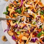 Asian Crunch Salad: a colorful salad with shredded cabbage, carrots, purple cabbage, green onions, cashews, crunchy noodles, and chopped herbs, drizzled with a brown dressing, served in a white bowl with a fork.