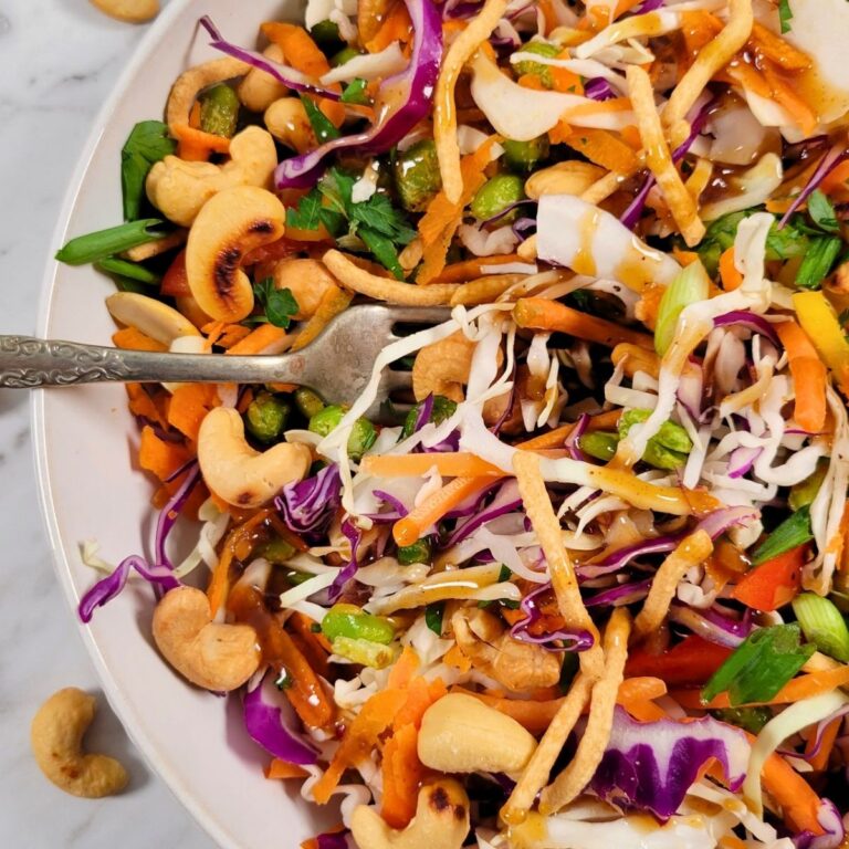 Asian Crunch Salad: a colorful salad with shredded cabbage, carrots, purple cabbage, green onions, cashews, crunchy noodles, and chopped herbs, drizzled with a brown dressing, served in a white bowl with a fork.