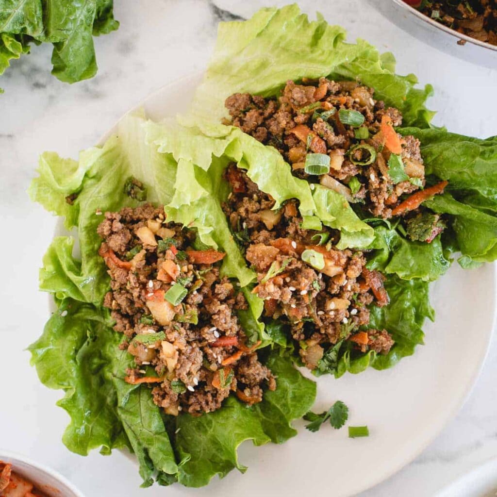 Lettuce leaves filled with seasoned ground meat, chopped vegetables, and garnished with sliced green onions, served on a white plate.