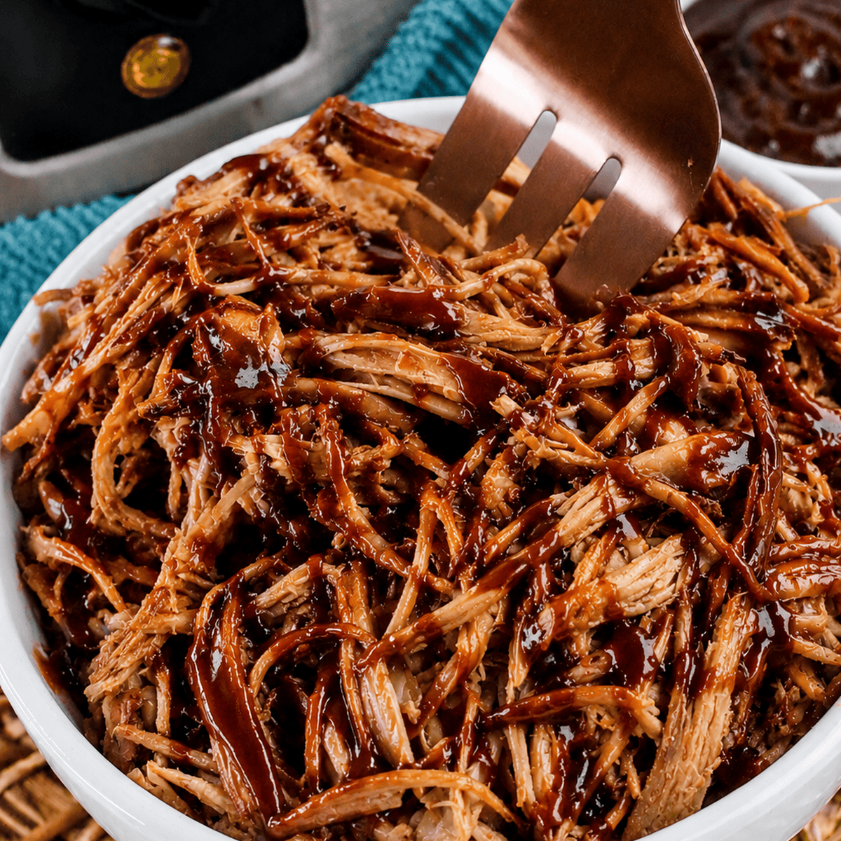 A close-up of a white bowl filled with Slow Cooker BBQ Pulled Pork, topped with barbecue sauce and being scooped with a large fork.