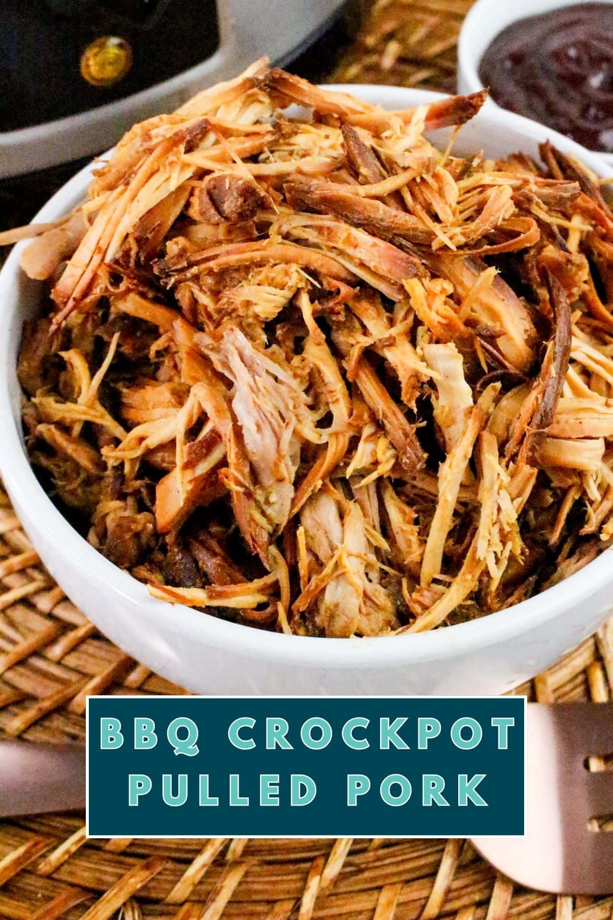 Slow Cooker BBQ Pulled Pork Recipe: a white bowl filled with shredded BBQ crockpot pulled pork sits on a woven mat, with a dish of barbecue sauce nearby. A text label at the bottom reads BBQ Crockpot Pulled Pork.