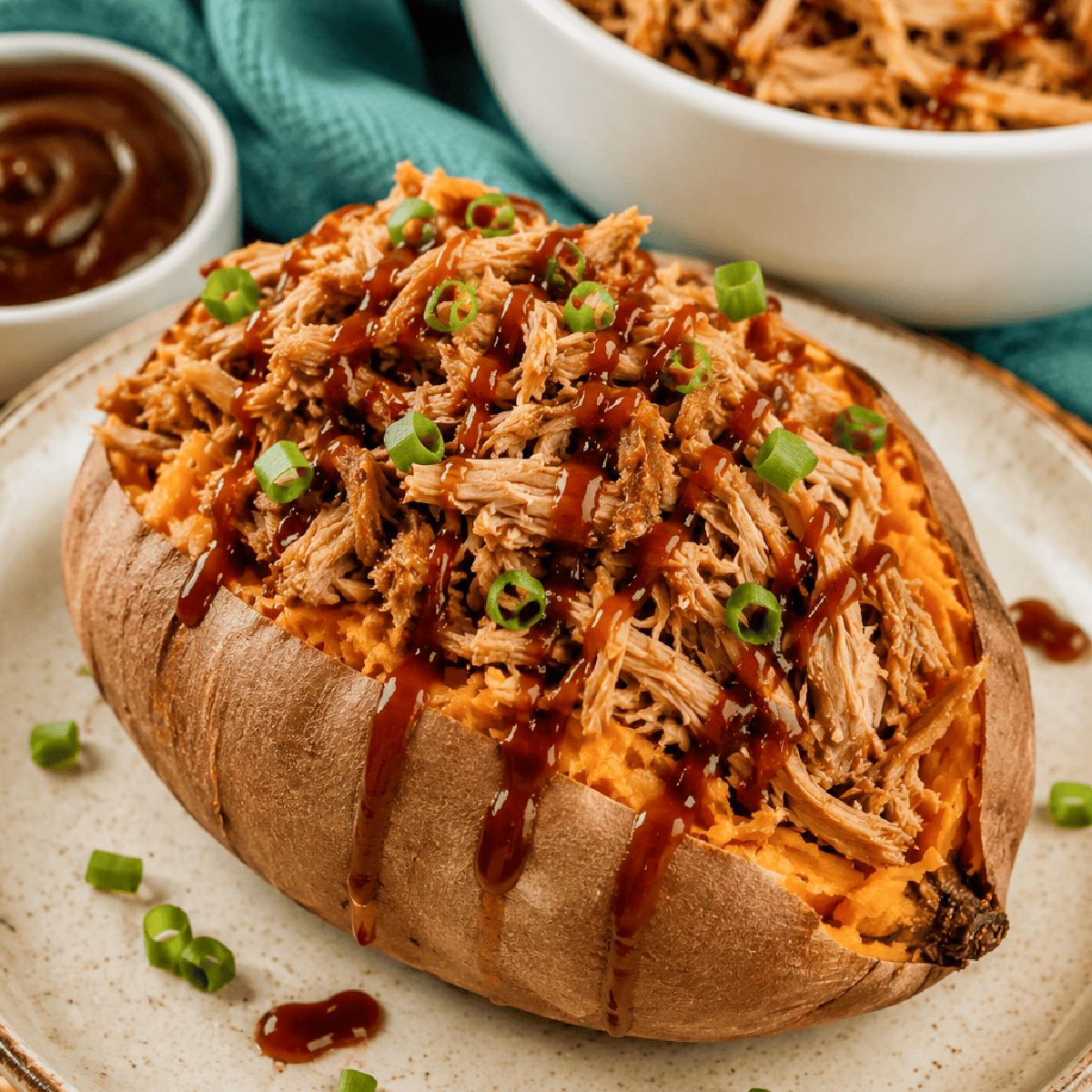 A baked sweet potato topped with Slow Cooker BBQ Pulled Pork, drizzled with barbecue sauce, and garnished with chopped green onions, served on a plate with a bowl of barbecue sauce in the background.