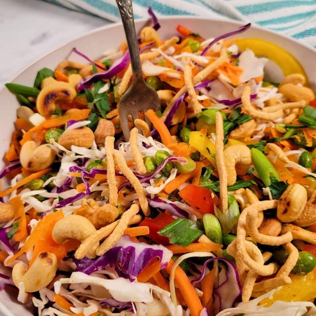 Crunchy Asian Salad: a vibrant salad with shredded cabbage, red cabbage, carrots, bell peppers, green onions, cashews, crunchy noodles, and herbs in a white bowl, with a fork resting inside.