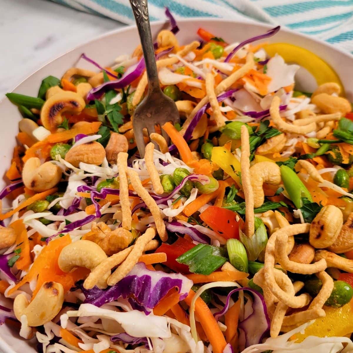 Crunchy Asian Salad: a vibrant salad with shredded cabbage, red cabbage, carrots, bell peppers, green onions, cashews, crunchy noodles, and herbs in a white bowl, with a fork resting inside.