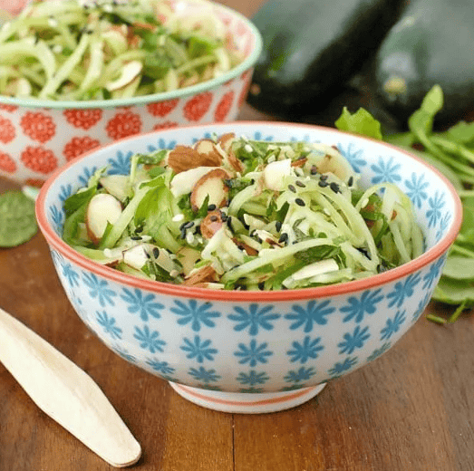 A bowl of fresh zucchini noodle salad garnished with sliced almonds and black sesame seeds sits on a wooden table. Another bowl of salad, zucchinis, and greens are visible in the background.