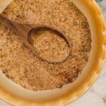 A wooden spoon rests in a shallow beige bowl filled with a coarse, mixed seasoning blend of various spices and herbs.