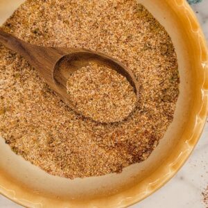 A wooden spoon rests in a shallow beige bowl filled with a coarse, mixed seasoning blend of various spices and herbs.