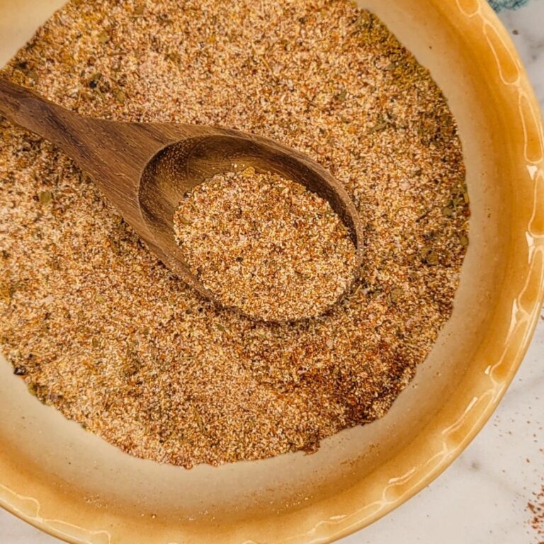 A wooden spoon rests in a shallow beige bowl filled with a coarse, mixed seasoning blend of various spices and herbs.