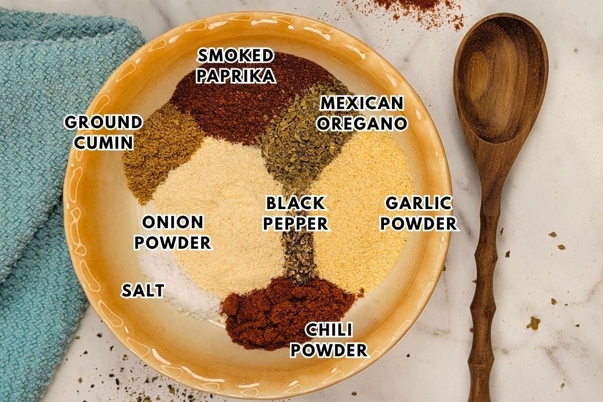 A bowl on a white surface contains labeled piles of spices: smoked paprika, ground cumin, Mexican oregano, black pepper, garlic powder, chili powder, onion powder, and salt. A wooden spoon and blue towel are nearby.