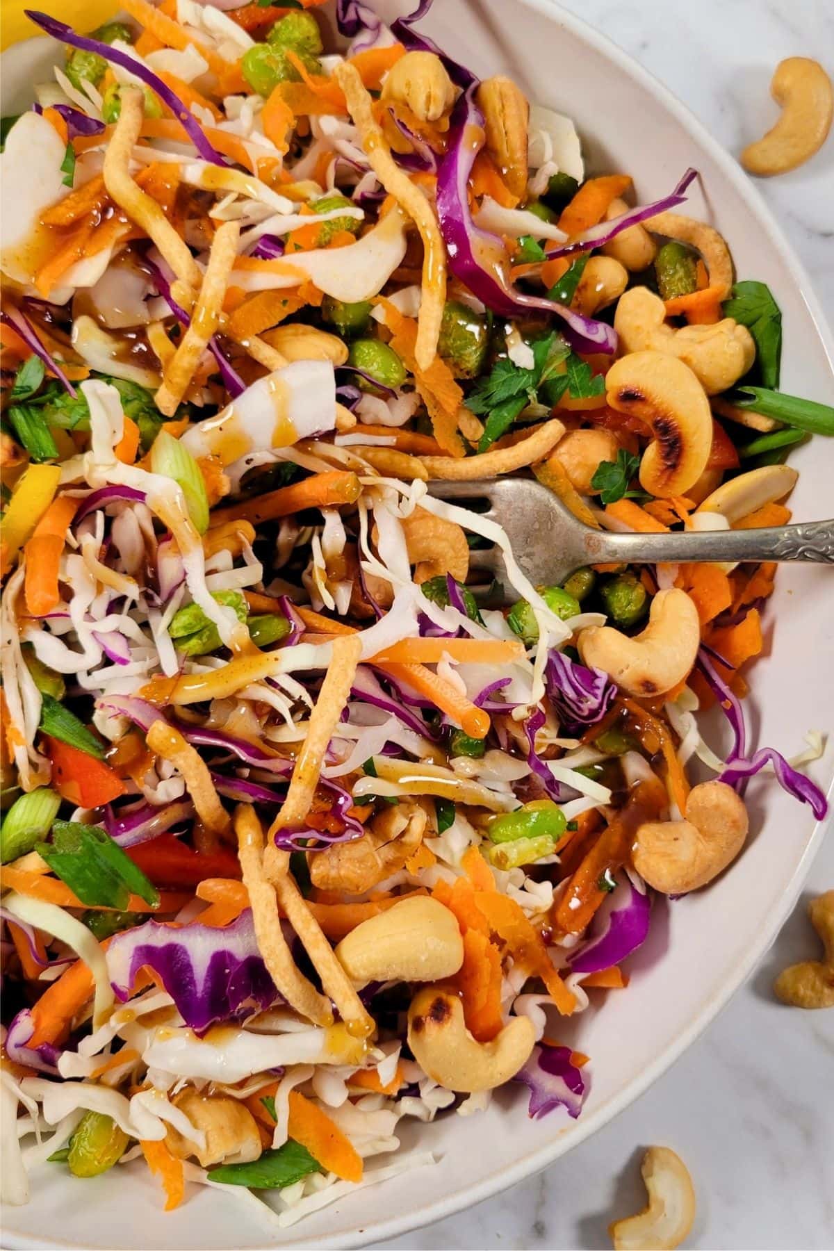 Asian Crunch Salad: a colorful salad in a white bowl with shredded cabbage, carrots, purple cabbage, green onions, cashews, cilantro, crispy noodles, and a brown dressing, with a fork resting inside.
