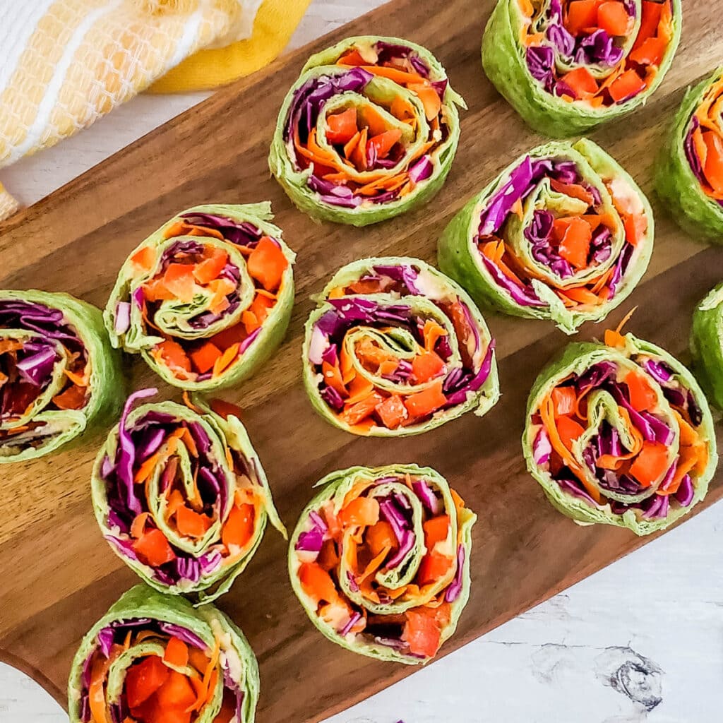 Colorful veggie pinwheels made with lettuce wraps, filled with shredded carrots, red cabbage, and red bell peppers, neatly arranged on a wooden serving board. A yellow and white towel is partially visible in the corner.
