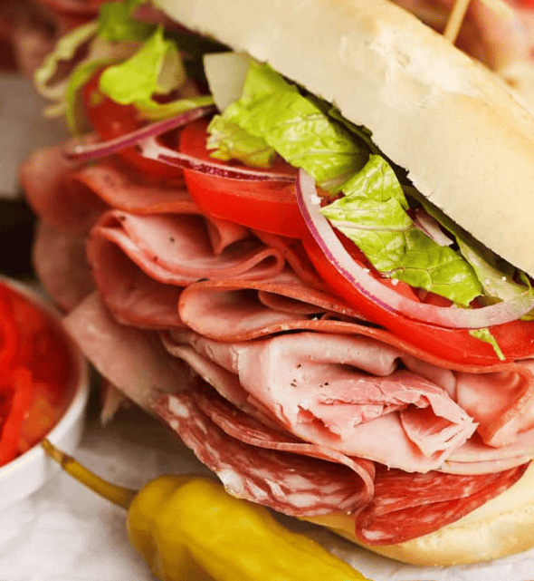 A close-up of an Italian sub sandwich filled with layers of cured meats, lettuce, tomato slices, red onion, and served with a pepperoncini and a side of sliced red peppers.