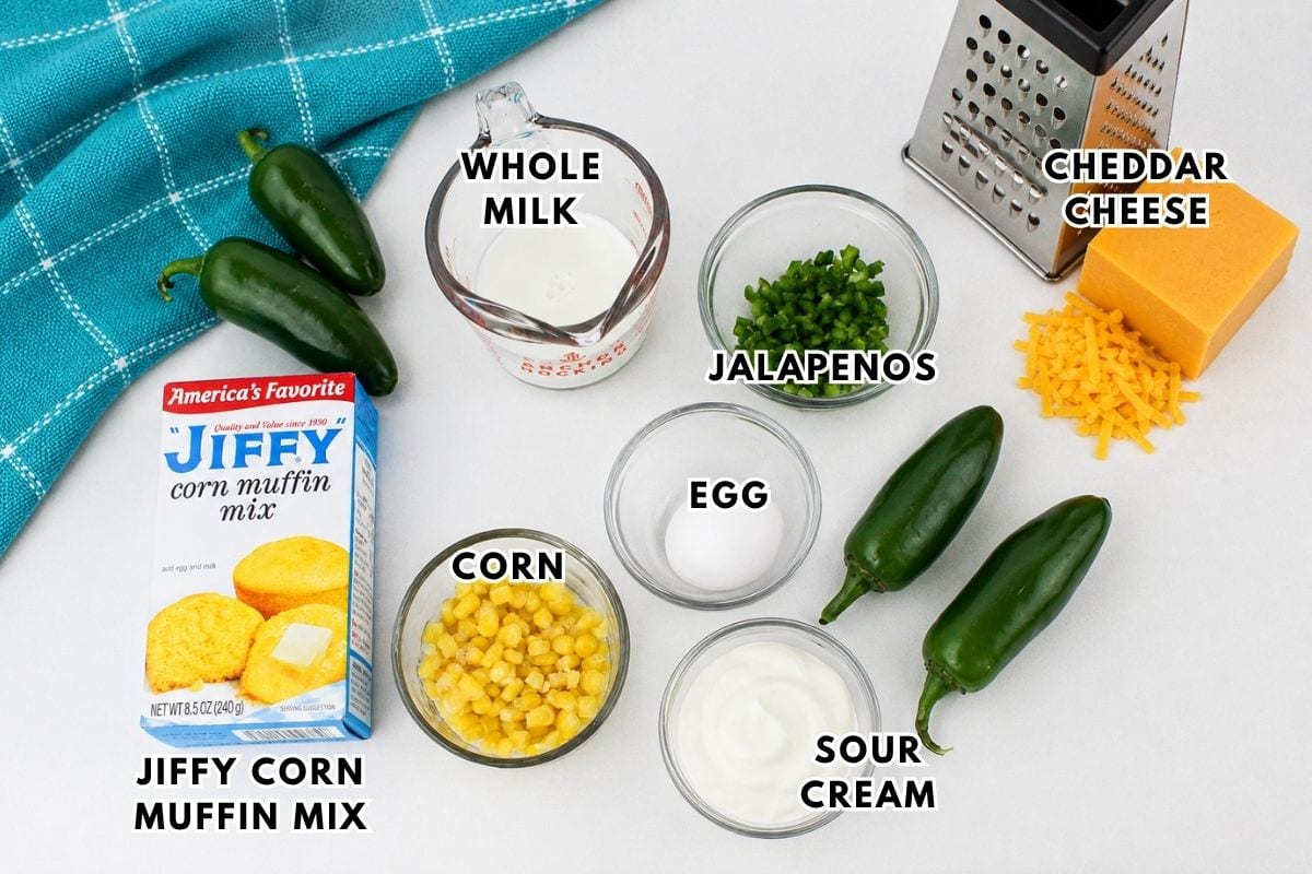 Ingredients for Jiffy Jalapeno Cornbread Muffin are arranged on a white surface: Jiffy corn muffin mix, whole milk, jalapeños, cheddar cheese, corn, egg, and sour cream. A grater and blue towel complete the setup.