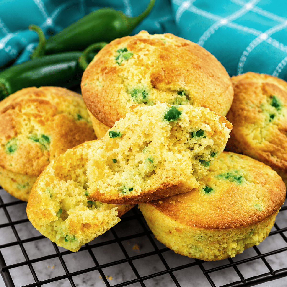 Jiffy Jalapeno Cornbread Muffins with green jalapeño pieces are stacked on a cooling rack, with one muffin split open to reveal the inside. Whole jalapeños and a blue checkered cloth sit in the background.