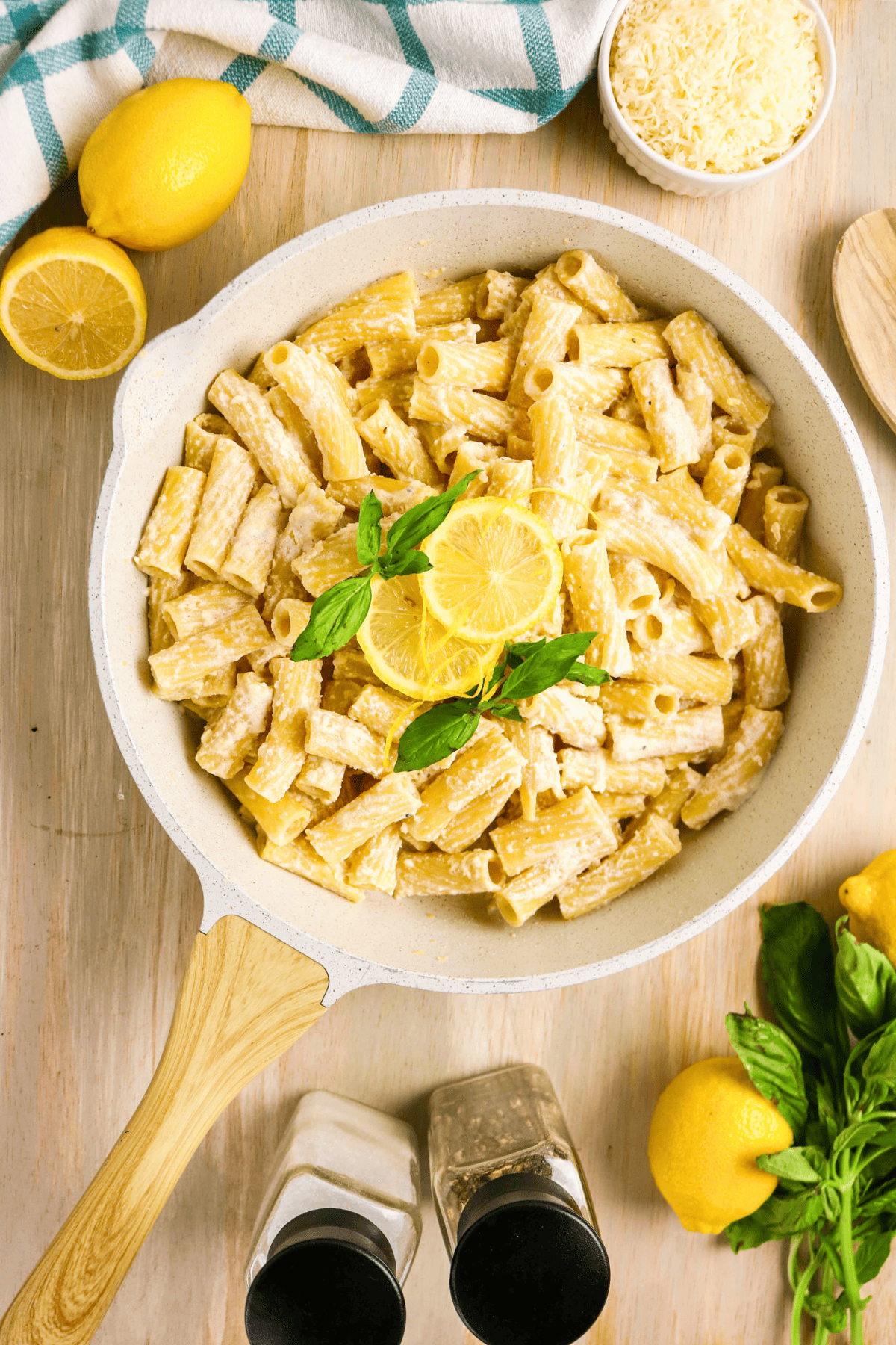 Pasta al Limone: a white skillet with creamy rigatoni pasta garnished with a lemon slice and fresh basil, surrounded by lemons, basil, grated cheese, pepper and salt shakers, and a blue-striped kitchen towel on a wooden surface.