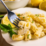 Lemon Ricotta Pasta: a fork holds creamy rigatoni pasta over a plate, garnished with chopped herbs and grated cheese. Lemon slices and fresh basil are visible on the plate beside the pasta.