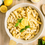 A skillet filled with creamy rigatoni pasta, garnished with fresh basil leaves and lemon slices, sits on a wooden surface with lemons and a small bowl of grated cheese nearby.