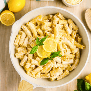 A skillet filled with creamy rigatoni pasta, garnished with fresh basil leaves and lemon slices, sits on a wooden surface with lemons and a small bowl of grated cheese nearby.