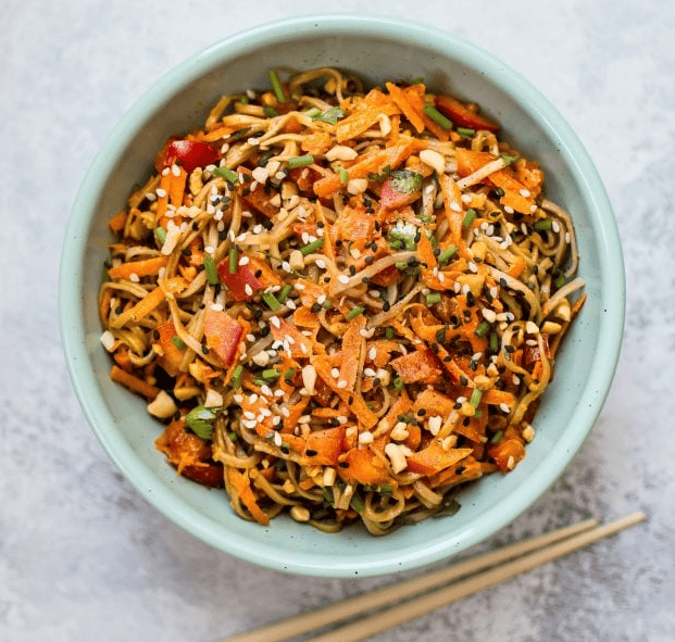 A bowl of colorful noodle salad with shredded carrots, red bell peppers, chopped herbs, and sesame seeds, served with chopsticks on a light surface.