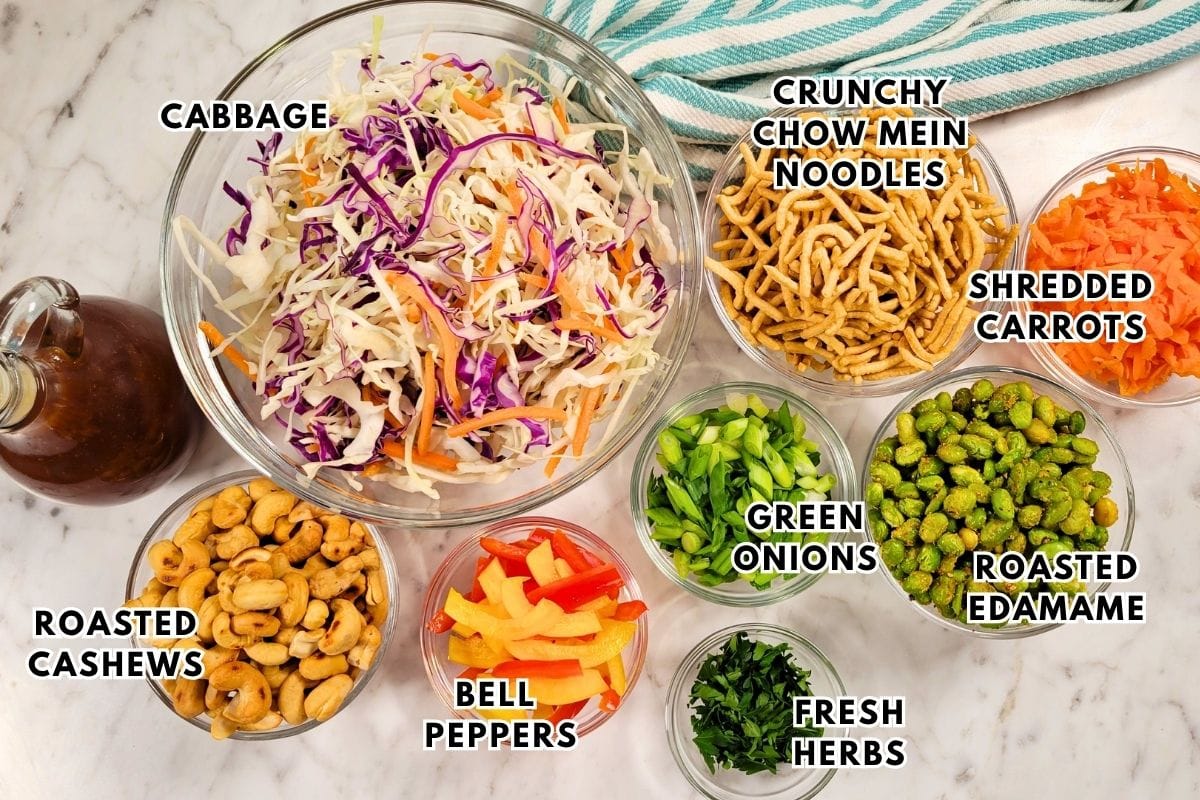 Cashew Crunch Salad: a variety of salad ingredients are in bowls on a white surface, labeled: cabbage, crunchy chow mein noodles, shredded carrots, green onions, roasted edamame, fresh herbs, bell peppers, roasted cashews, and dressing.