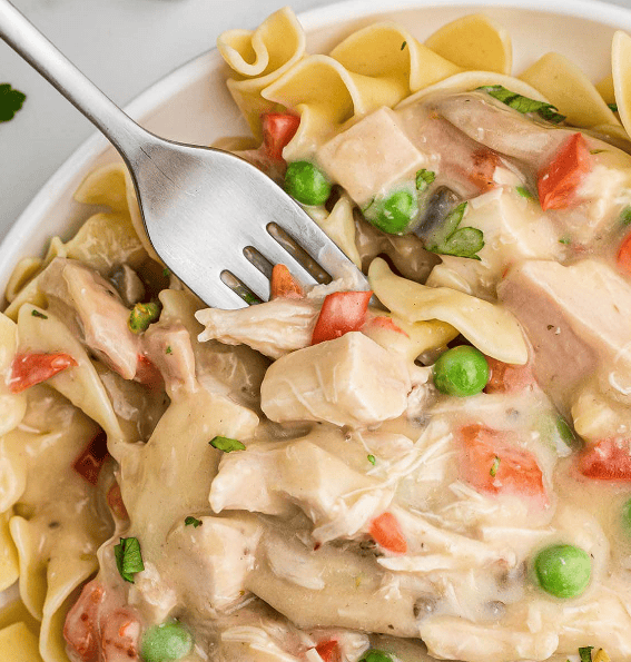 A close-up of creamy chicken and noodles with peas and diced red peppers, served on a white plate. A fork lifts a bite, highlighting tender chicken and wide egg noodles—an easy favorite from 30 Minute Chicken Recipes.