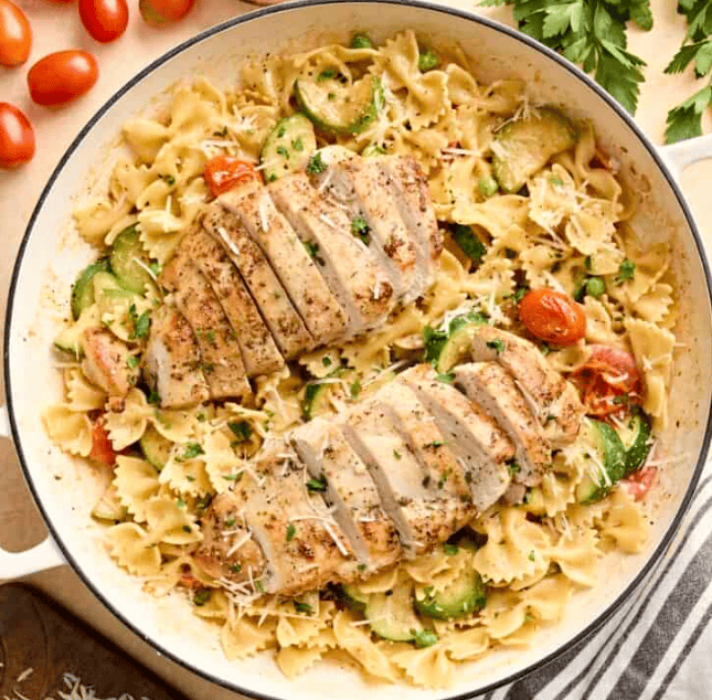 A skillet filled with bow-tie pasta, sliced grilled chicken breasts, zucchini, cherry tomatoes, and grated cheese—one of the best 30 Minute Chicken Recipes. Fresh parsley and tomatoes are placed nearby for a vibrant finishing touch.