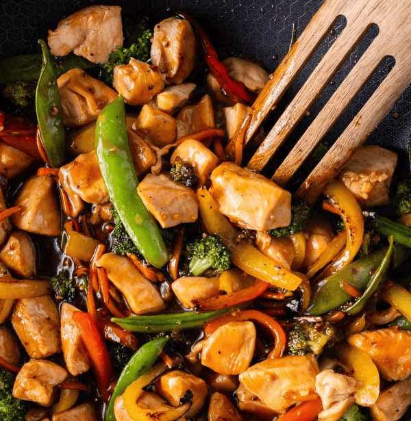 A close-up of a wooden spoon stirring a colorful chicken stir-fry with snow peas, bell peppers, carrots, broccoli, and sauce in a pan—perfect for quick 30 Minute Chicken Recipes.