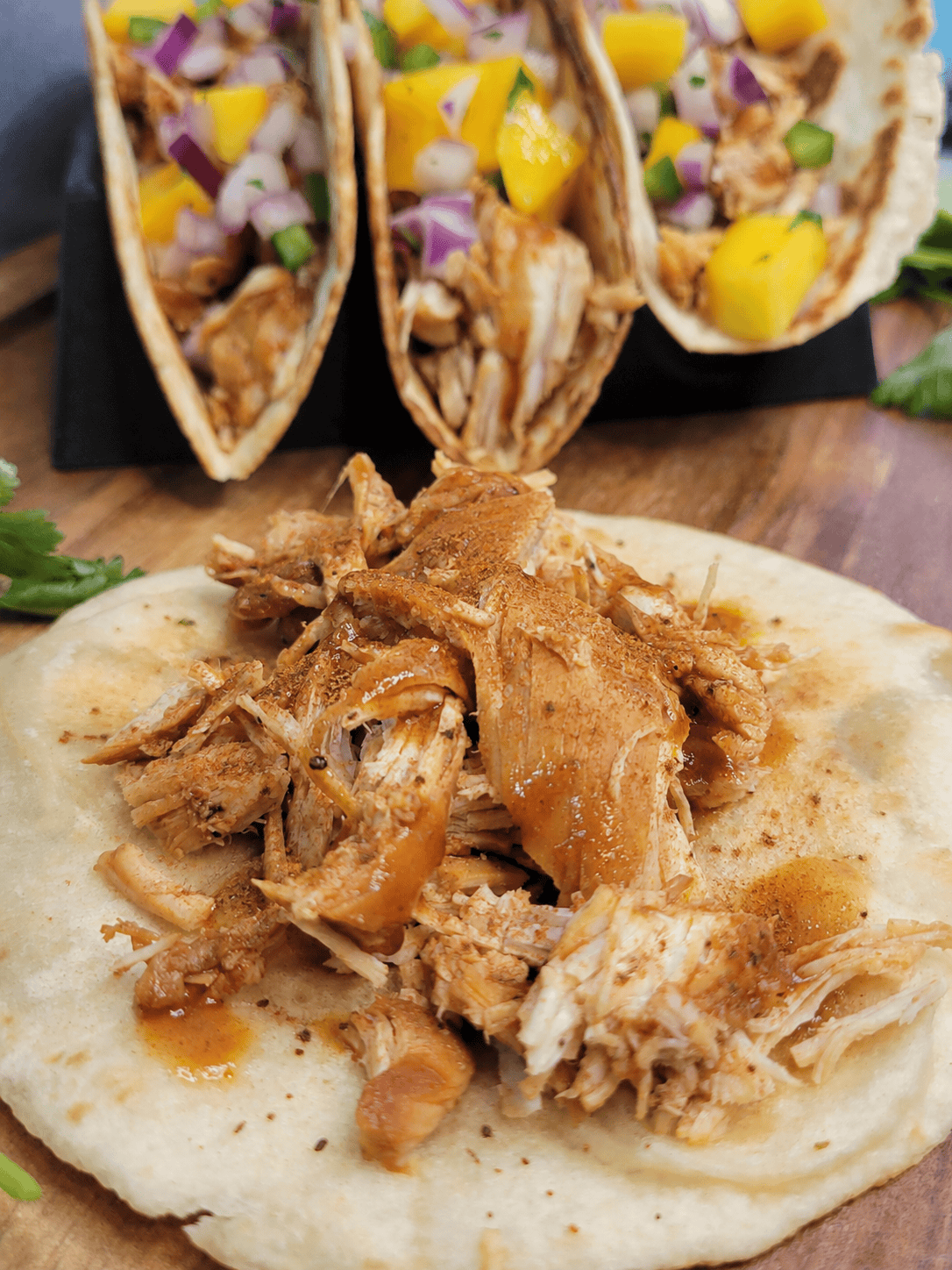Shredded chicken tossed in Chicken Taco Seasoning sits on a tortilla in the foreground, while two tacos filled with chicken, diced mango, red onion, and peppers are arranged on a wooden surface in the background.