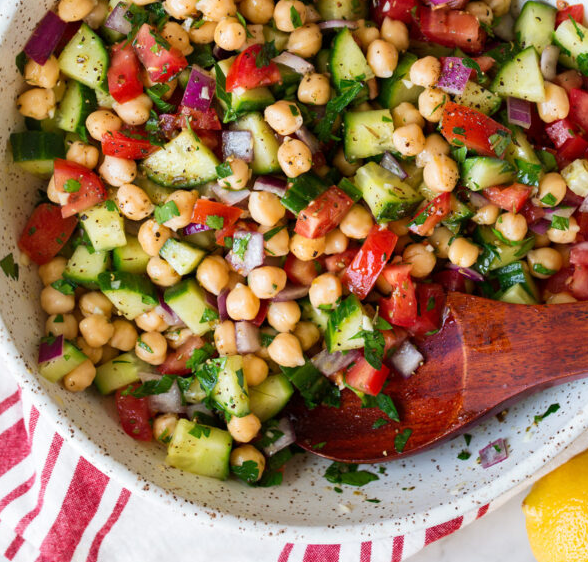 A close-up of a colorful chickpea salad with diced cucumbers, tomatoes, red onions, parsley, and seasoning, served in a bowl with a wooden spoon and a striped red and white napkin nearby.