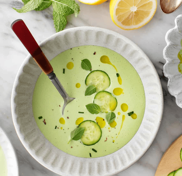 A bowl of creamy green soup garnished with cucumber slices, fresh herbs, a drizzle of oil, and cracked pepper. A spoon rests in the bowl; lemon halves and mint leaves are nearby on a marble surface.