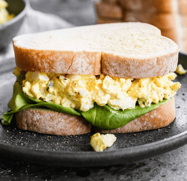 Egg salad sandwich with leafy greens on thick slices of white bread, served on a dark plate with a small piece of filling fallen beside it.