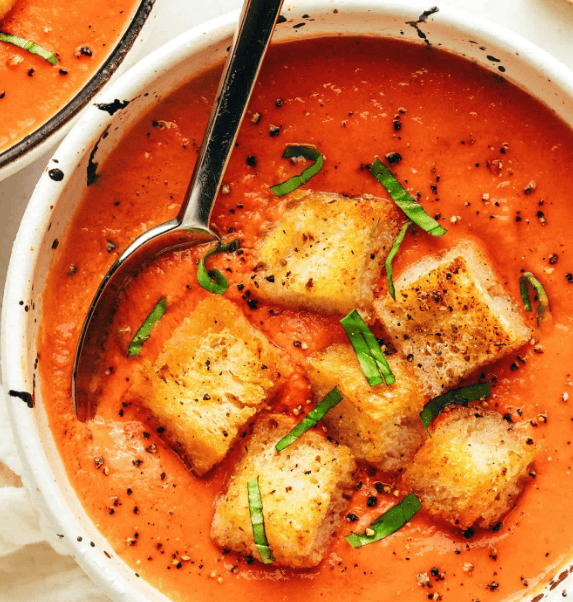 A bowl of tomato soup topped with crispy croutons, black pepper, and fresh basil strips, with a spoon resting inside the bowl.