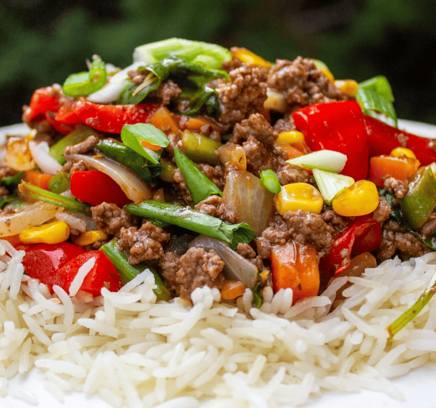 A close-up of a plate of white rice topped with cooked ground beef, sliced red bell peppers, corn, chopped green onions, and other mixed vegetables.