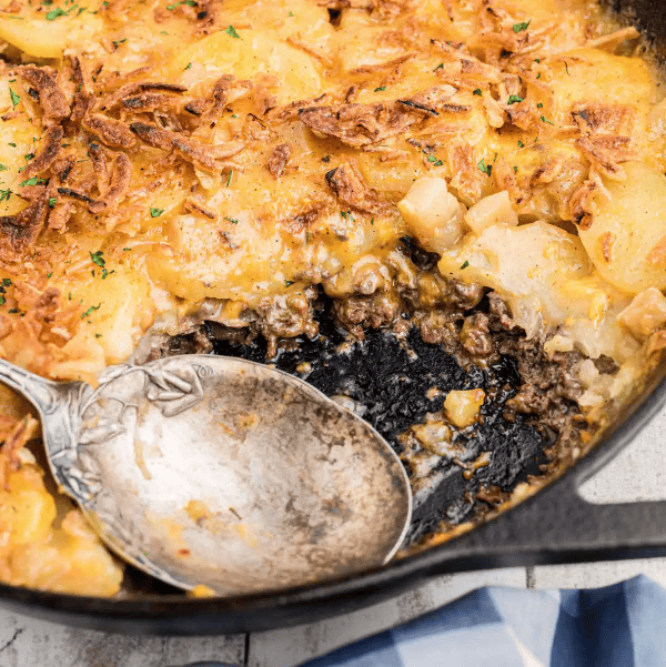 A close-up of a baked casserole in a skillet, topped with golden brown potato slices and crispy onions. A large serving spoon rests inside, revealing layers of ground beef and creamy sauce underneath.