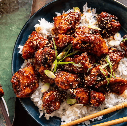 A plate of white rice topped with glazed, sesame-seed sprinkled chicken pieces, garnished with sliced green onions—perfect for those seeking quick and tasty 30 Minute Chicken Recipes. Wooden chopsticks rest beside the food.