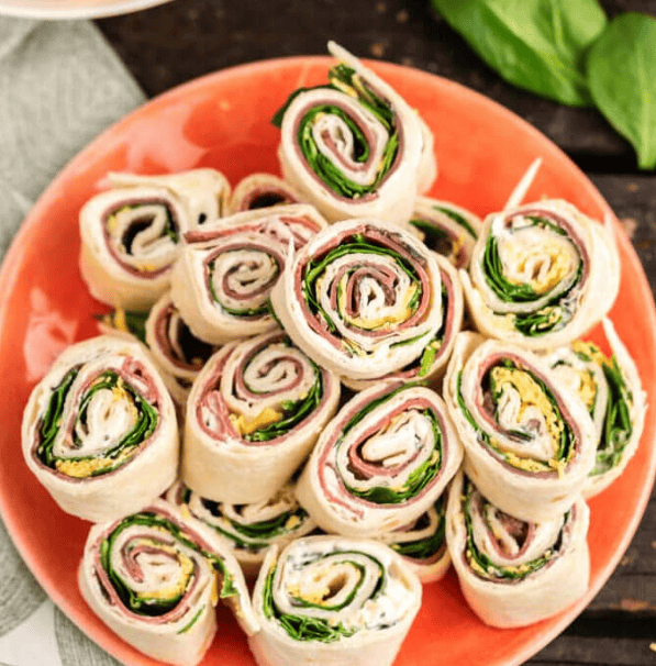 A stack of tortilla pinwheels filled with deli meat, cheese, cream cheese, and spinach, arranged on an orange plate on a wooden surface.