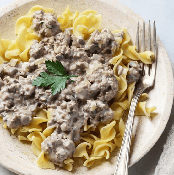 A plate of egg noodles topped with creamy ground beef stroganoff, garnished with a sprig of parsley, next to a fork on a light-colored plate.