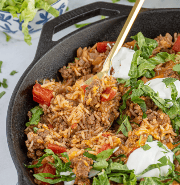 A close-up of a skillet filled with taco rice, ground beef, diced tomatoes, melted cheese, shredded lettuce, and dollops of sour cream, with a spoon scooping up a portion.