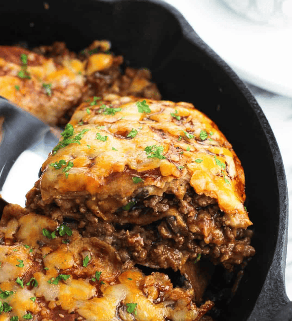 A close-up of a cheesy, baked casserole with layers of ground beef and sauce, topped with melted cheese and garnished with chopped herbs, served in a black skillet.