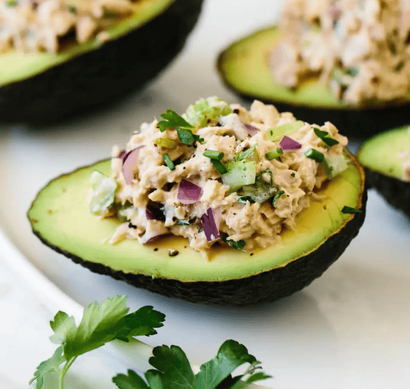 Halved avocado filled with a creamy mixture of tuna, chopped red onion, celery, and herbs, garnished with parsley, served on a white plate.