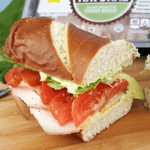 A close-up of a sandwich on a wooden surface, filled with sliced turkey, tomato, lettuce, and cheese on a pretzel bun. Packaged deli meat is blurred in the background.