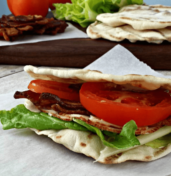 A BLT sandwich with bacon, lettuce, tomato, and mayonnaise on flatbread sits on parchment paper, with more flatbreads, bacon, lettuce, and tomato in the background.