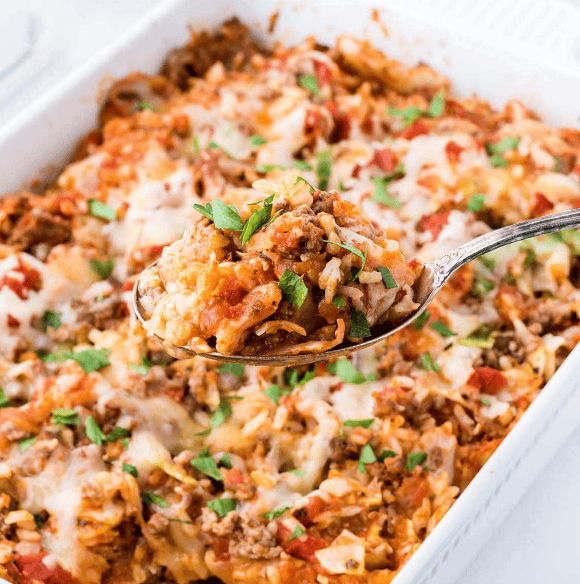 A close-up of a spoonful of cheesy, baked casserole with ground meat, rice, tomatoes, and sprinkled fresh herbs—one of the best dinner ideas with ground beef—held above a white baking dish filled with the same casserole.